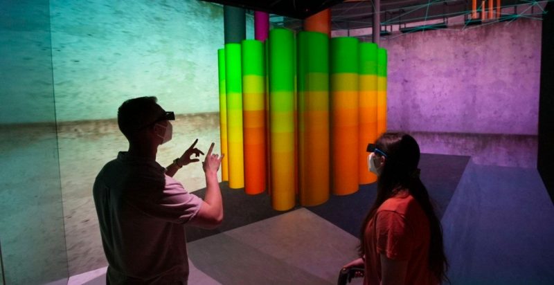 Two people with VR goggles on look at colorful cylinders.